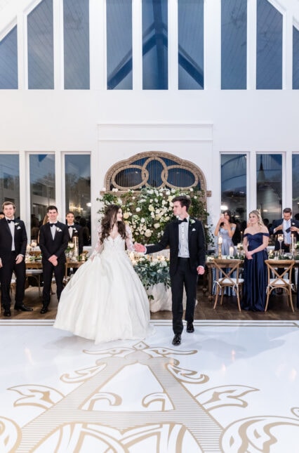 Bride and groom sharing their first dance on a custom gold monogram dance floor inside The French Farmhouse Venue reception hall with navy bridesmaid dresses and candlelit tables