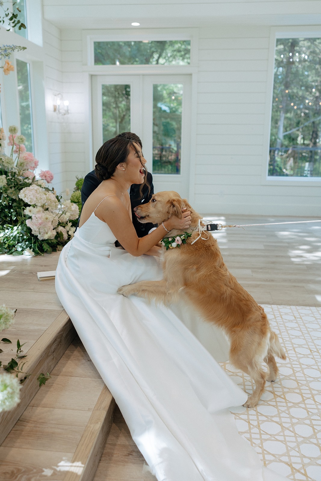 Ally laughing as their golden retriever wearing a pink floral collar jumps up on her lap at the chapel altar steps, Drew smiling beside her, with the floral arch and windows behind them