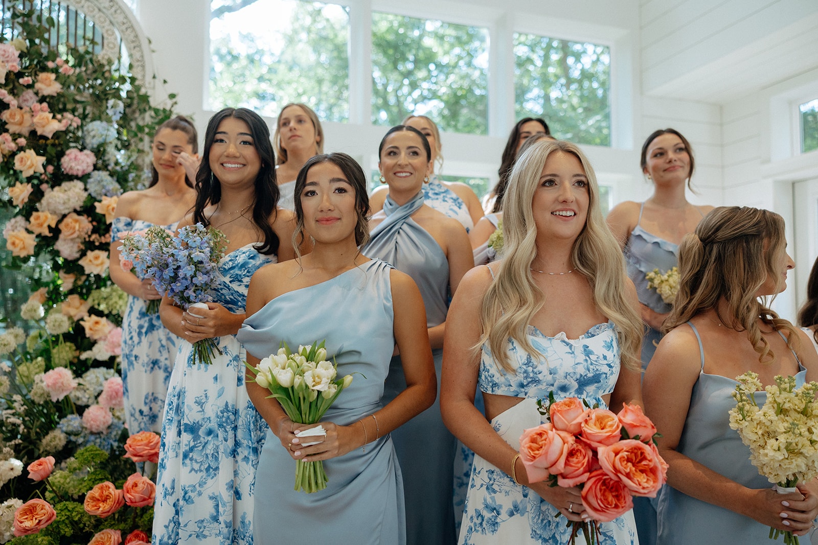 Ally's bridesmaids in a mix of blue and white floral print gowns and solid dusty blue satin dresses in various styles — one-shoulder, halter, spaghetti strap, cutout — holding bouquets of blue delphinium, white tulips, and coral garden roses, standing beside the floral arch with tears in their eyes watching the ceremony