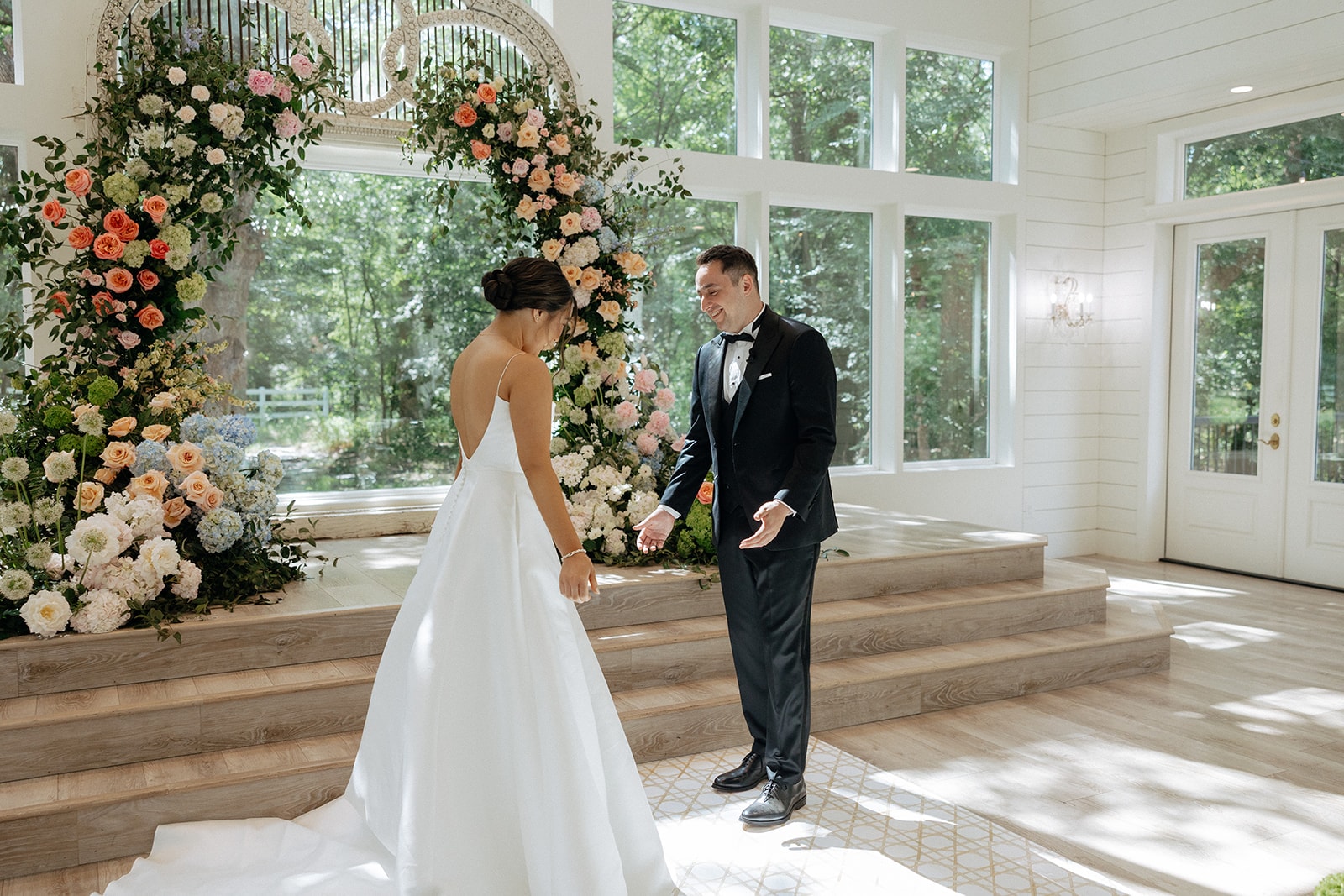 Wide shot of The French Farmhouse chapel ceremony setup — soaring cathedral ceilings with exposed wood beams and gold chandeliers, white Chiavari chairs lining the aisle, wicker baskets overflowing with blue, white, coral, and green hydrangeas on the floor, and the colorful floral arch at the altar
