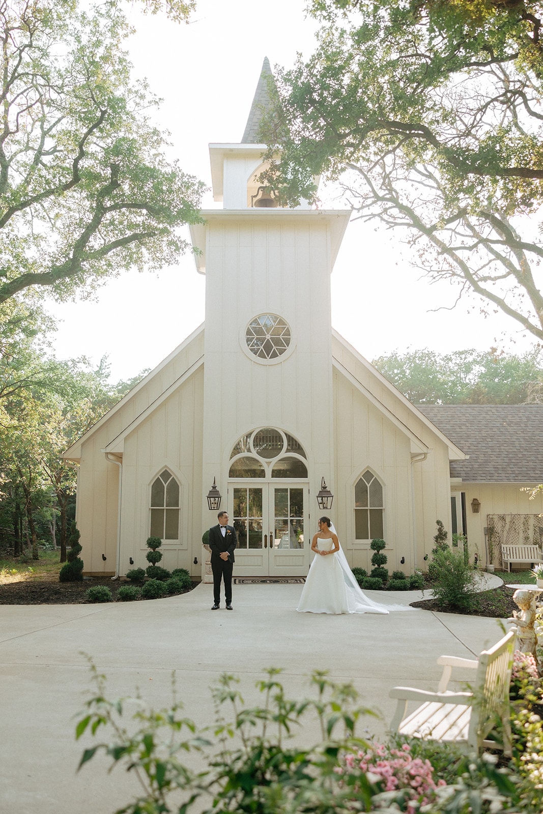 Ally and Drew standing apart in front of the white chapel exterior — Drew in a black tuxedo, Ally in her strapless gown with cathedral train — surrounded by mature oak trees, pink roses, and the chapel's gothic arched windows and steeple