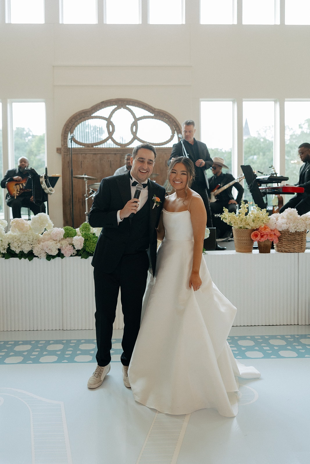 Drew singing into a microphone with his arm around Ally on the reception dance floor, the Royal Dukes Band performing behind them on a white fluted stage, low garden arrangements of hydrangeas and coral roses lining the stage front