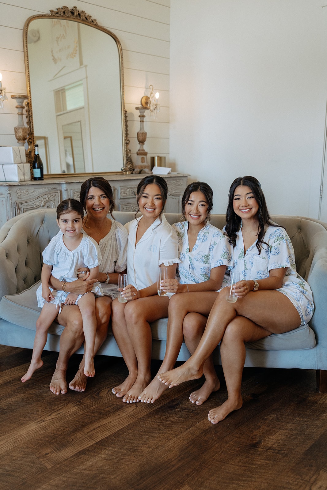 Ally with her mother, sister, and two bridesmaids in matching blue hydrangea floral pajama sets and a white ruffle set, sitting on the tufted sage velvet sofa in the bridal suite holding champagne flutes, with the ornate gold mirror and antique mantle behind them