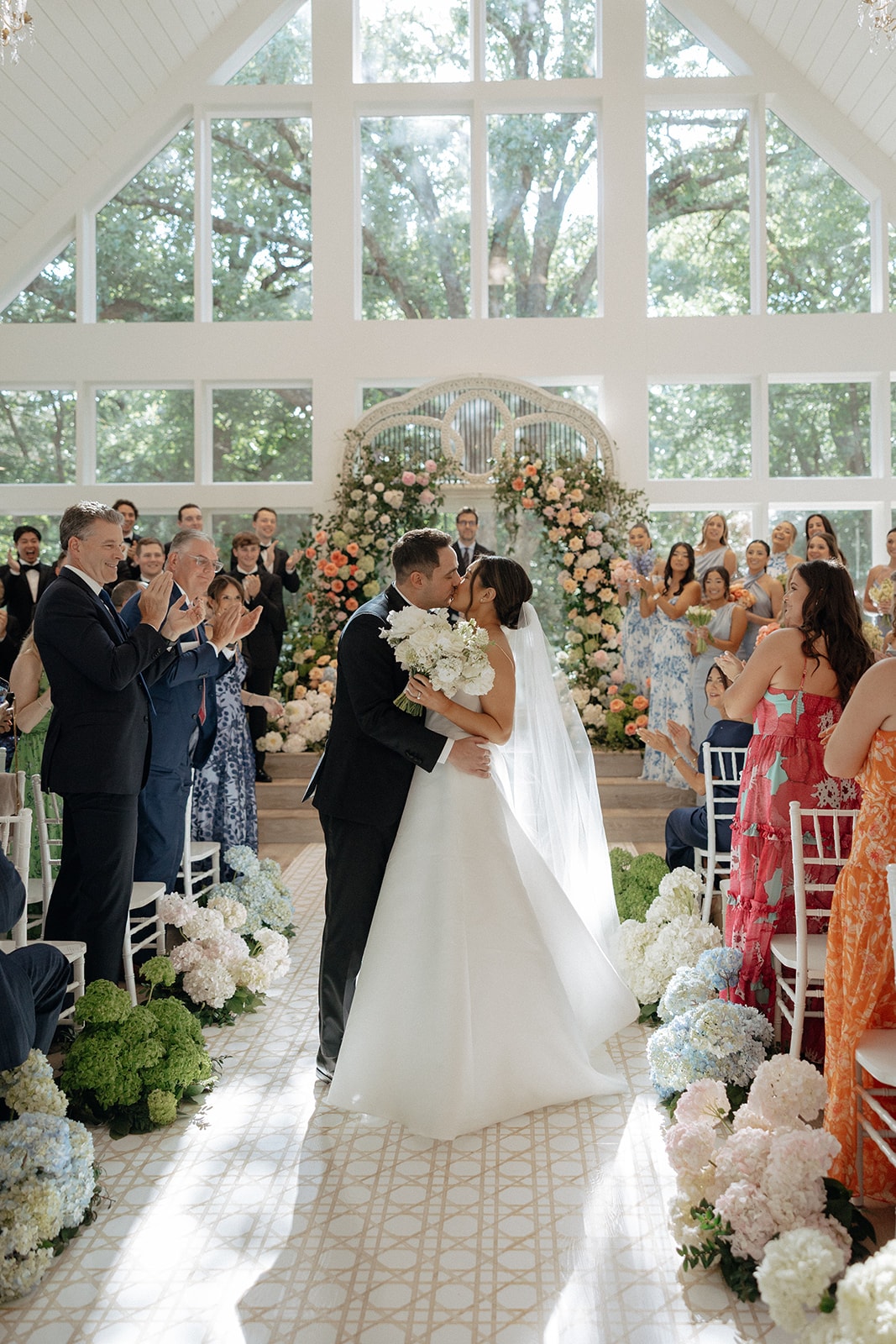 Aerial view of the reception hall with long farm tables in white lace and dusty blue linen, cane-back bistro chairs, low garden centerpieces of hydrangeas and coral roses, taper candles, and scattered citrus fruit, the wedding cake on the mantle in the background