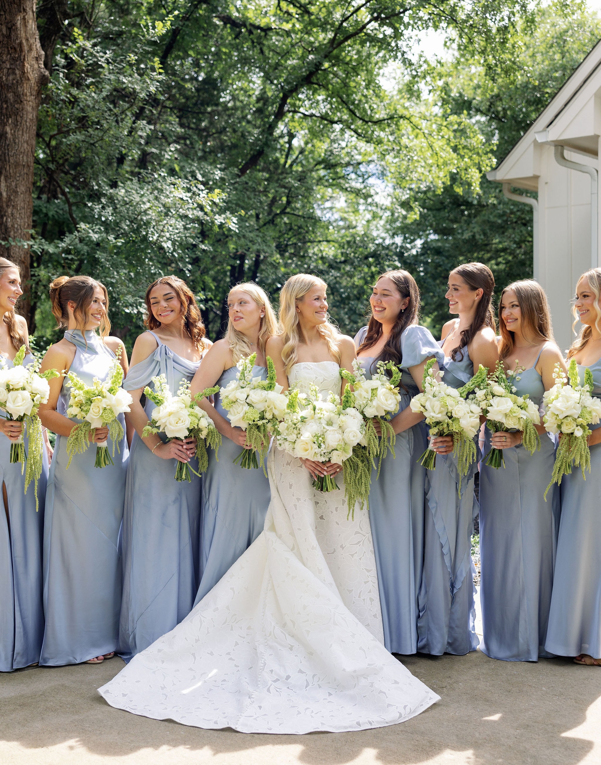 Bride and bridesmaids in dusty blue gowns holding white and green bouquets outdoors