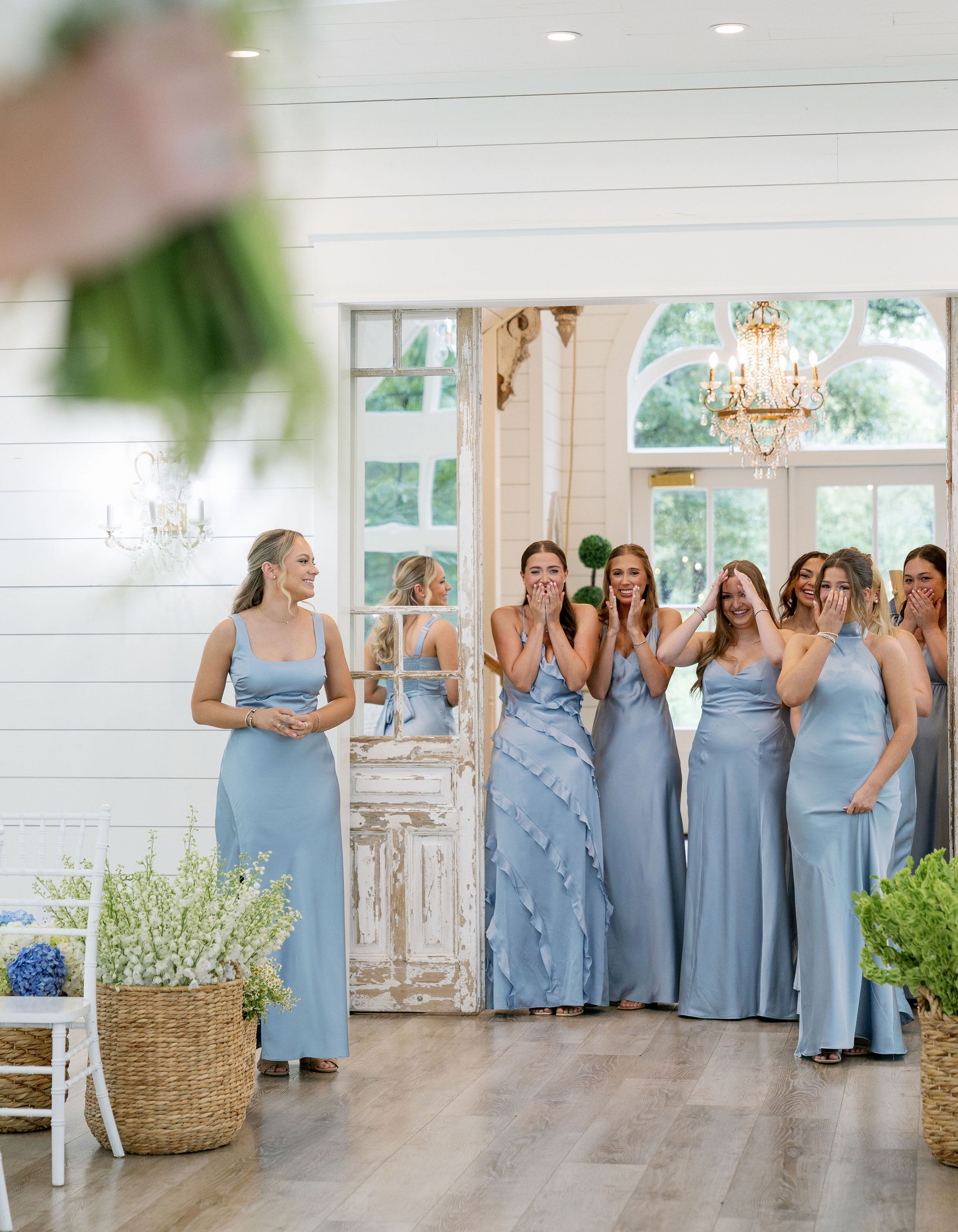 Bridesmaids in dusty blue gowns reacting with joy at the bride reveal