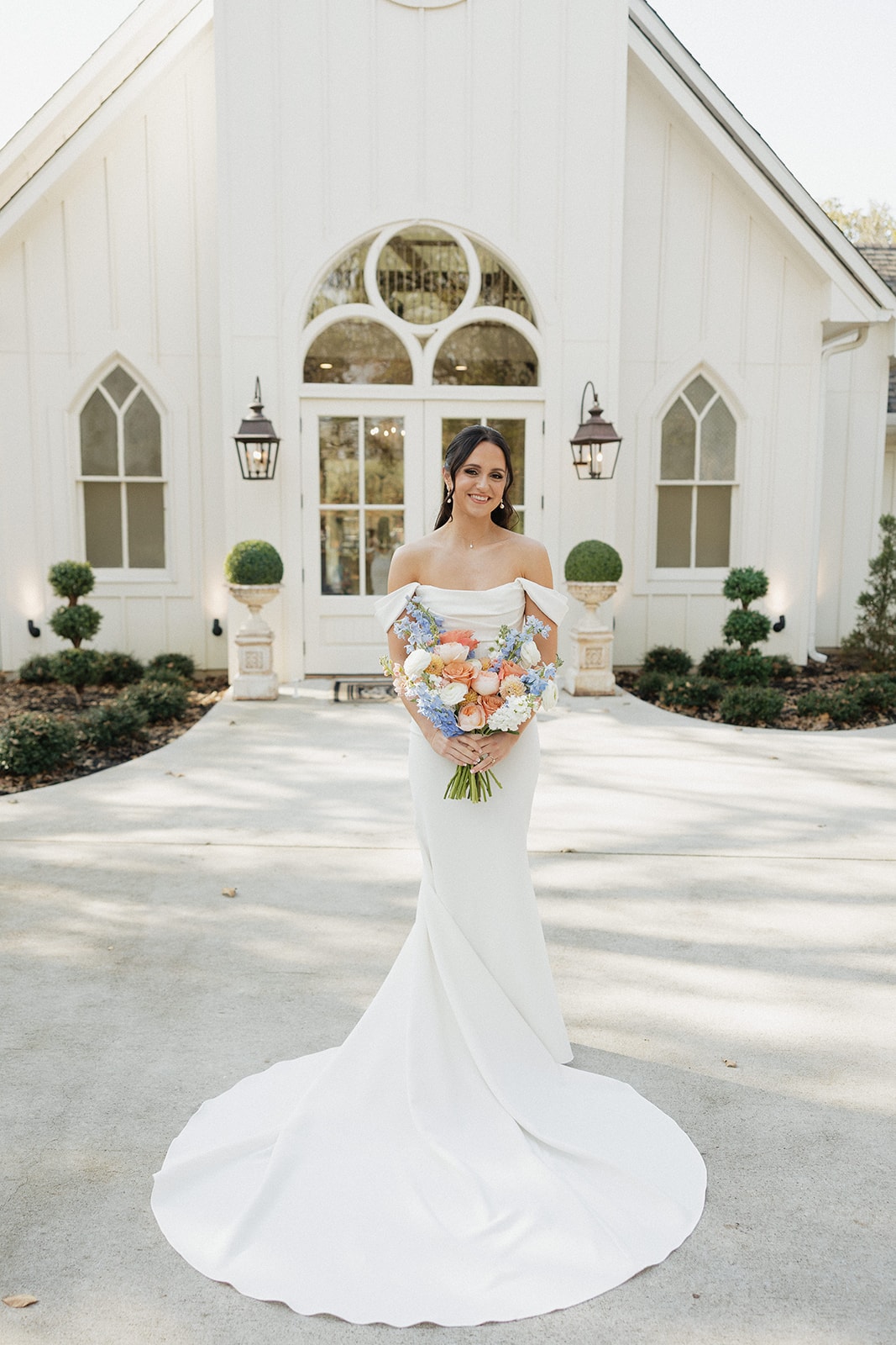 Avery standing in front of the chapel in her off-shoulder gown holding a coral and blue bouquet
