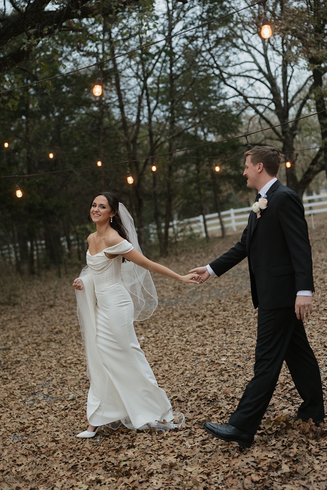 Avery and Pearson walking hand in hand under string lights at dusk