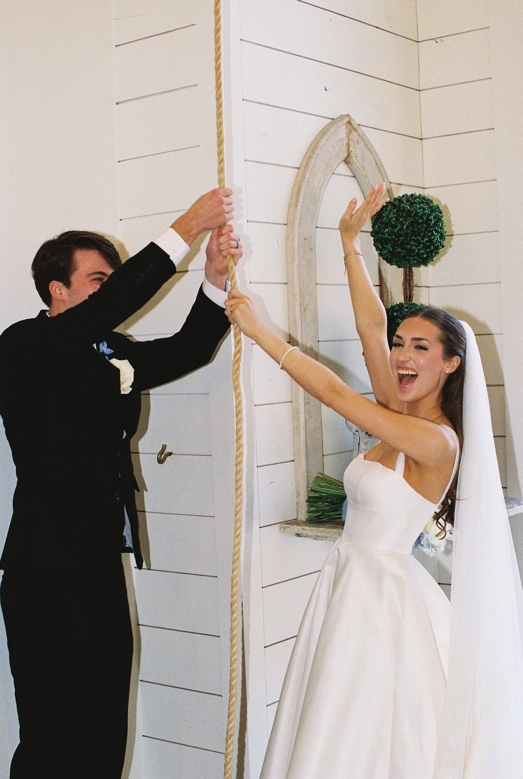 Brooke throwing her hands up in pure joy as she and Shepard pull the rope to ring the French Farmhouse chapel bell together, both laughing, the white shiplap wall and antique arch behind them
