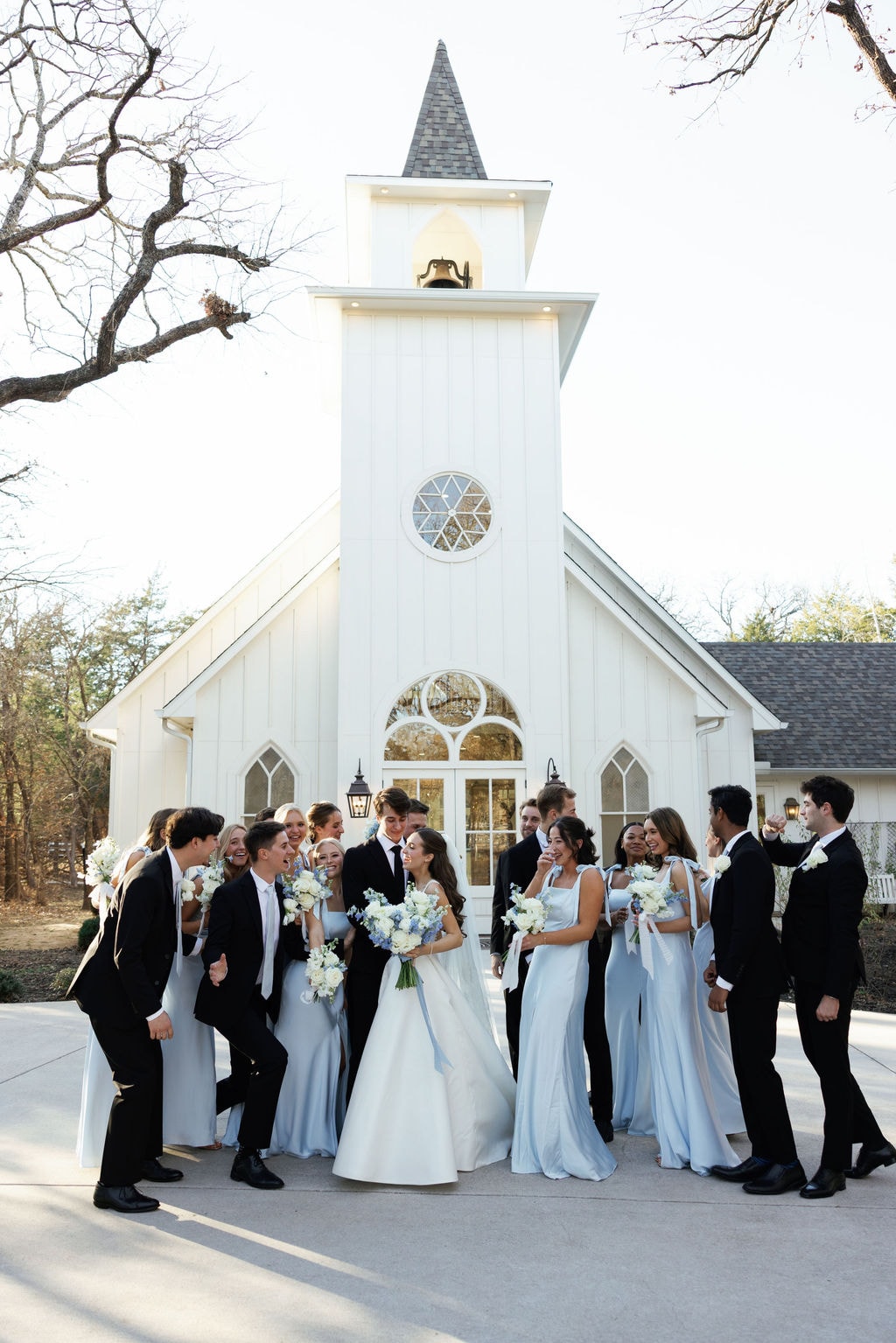 Brooke and Shepard surrounded by their full bridal party in ice blue dresses and black tuxedos, laughing together in front of the white French Farmhouse chapel steeple