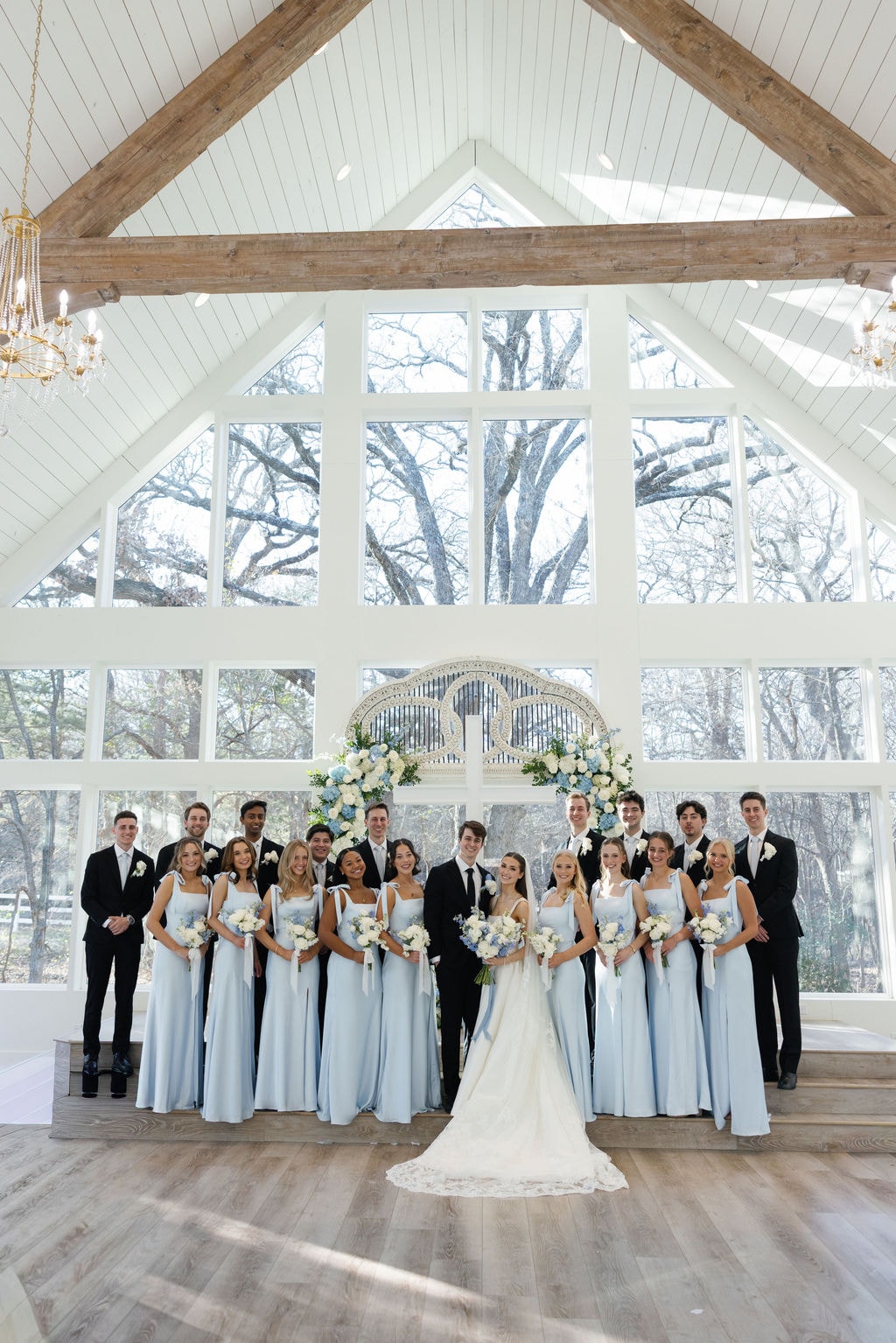 Full bridal party portrait inside the French Farmhouse chapel — ice blue bridesmaids and black tuxedos flanking Brooke and Shepard in front of the antique arch and floor-to-ceiling windows with bare winter oak trees beyond