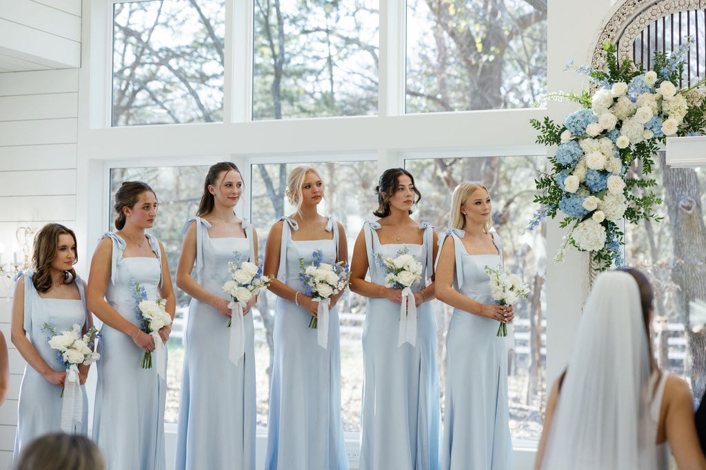 Six bridesmaids in ice blue tie-shoulder gowns holding white and blue bouquets, standing in front of the floor-to-ceiling chapel windows with the blue hydrangea and white rose floral arch visible to the right
