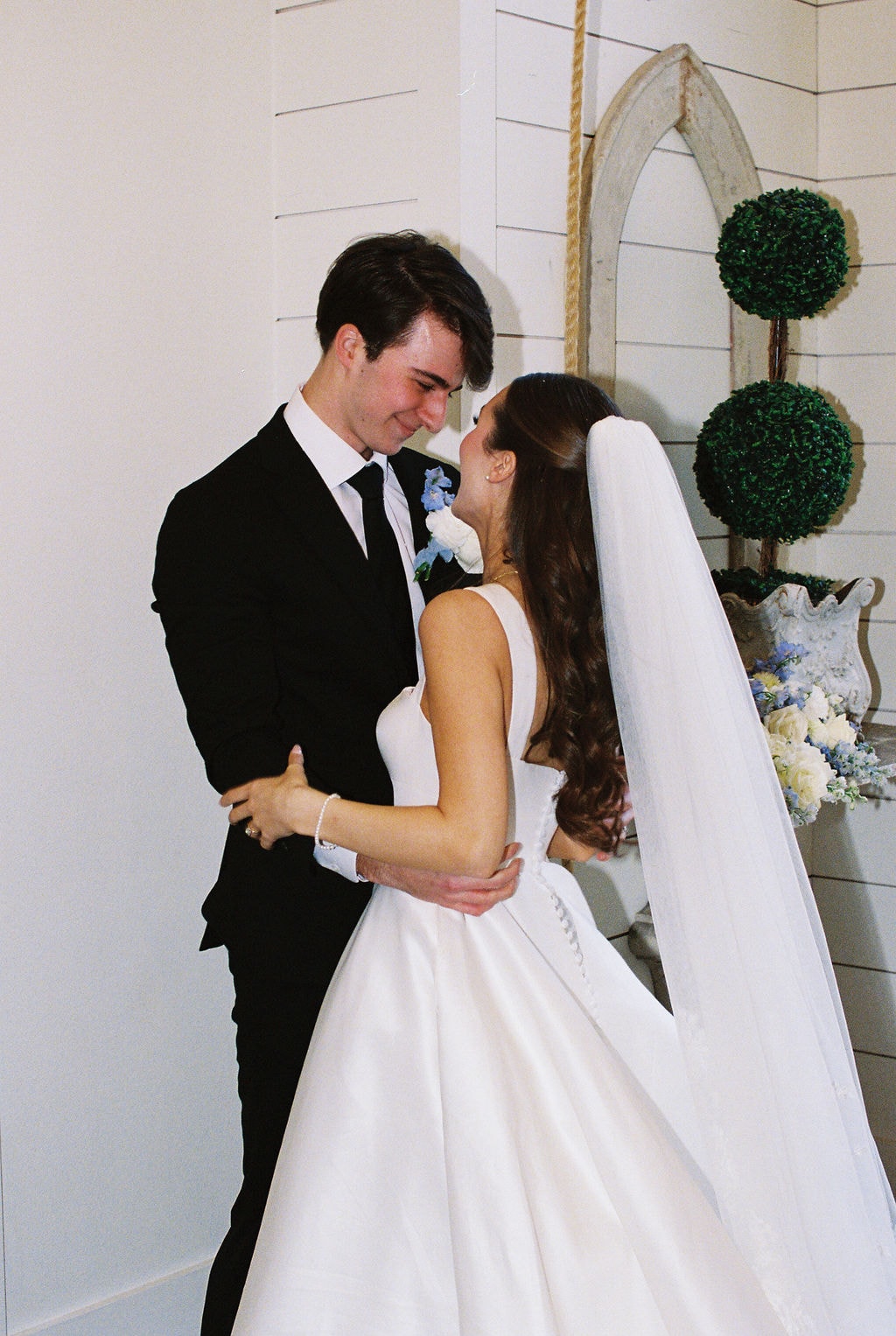 Brooke and Shepard gazing at each other in a tender moment beside the antique arch doorway, Brooke's veil trailing behind her, blue delphinium and white roses in the background
