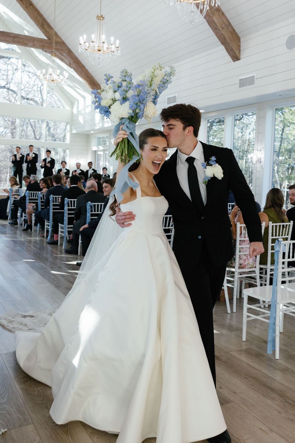 Brooke and Shepard holding hands at the altar during their vows, the ornate white antique arch flanked by cascading blue hydrangea and white rose arrangements, floor-to-ceiling windows with bare winter oak trees behind them
