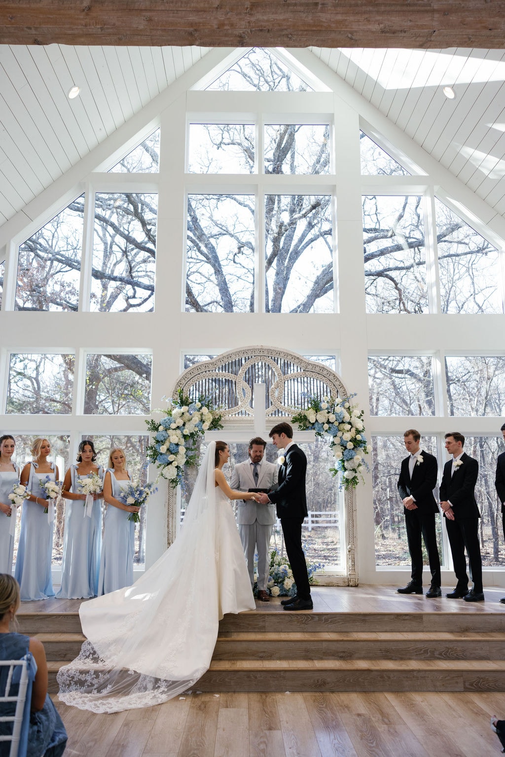 Wide shot of Brooke and Shepard's ceremony — the full chapel with ice blue chair sashes, bridesmaids and groomsmen flanking the couple at the altar beneath the soaring cathedral windows and blue hydrangea arch