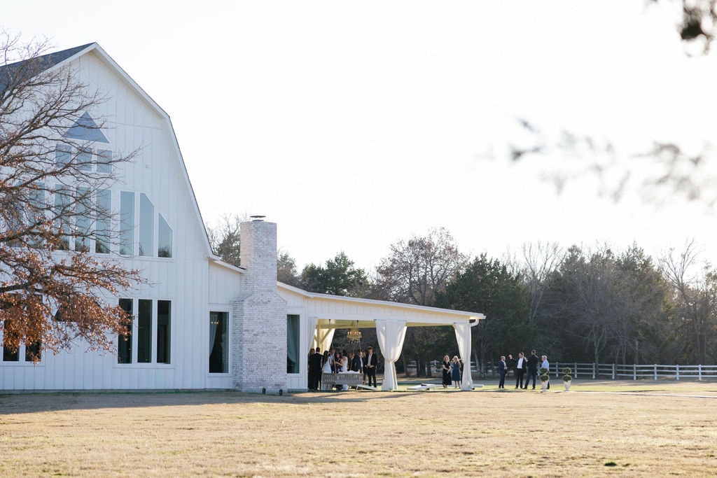 Wide shot of guests mingling under the white pergola pavilion with chandelier and draping curtains beside the French Farmhouse reception barn, bare winter oak trees and white fence in the background on a clear January afternoon