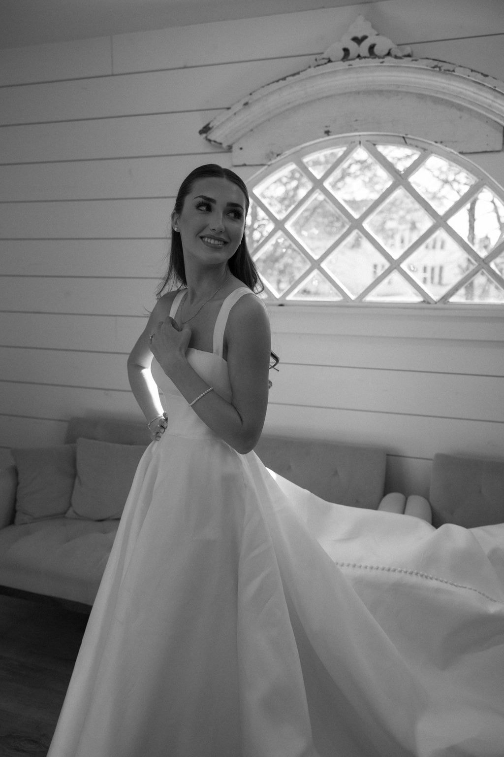 Black and white portrait of Brooke in her structured square-neck ballgown in the mini bridal suite, smiling over her shoulder in front of the arched diamond-pane window and white shiplap walls