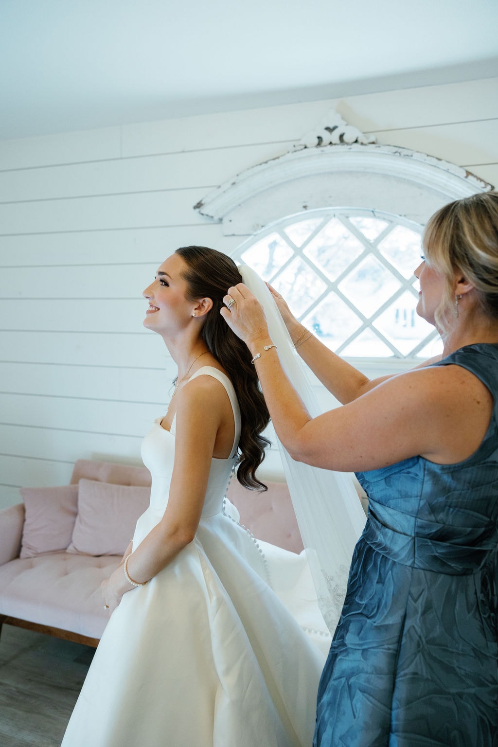 Brooke beaming as someone places her veil in the mini bridal suite, the arched diamond-pane window and blush pink sofa of the French Farmhouse chapel suite behind them