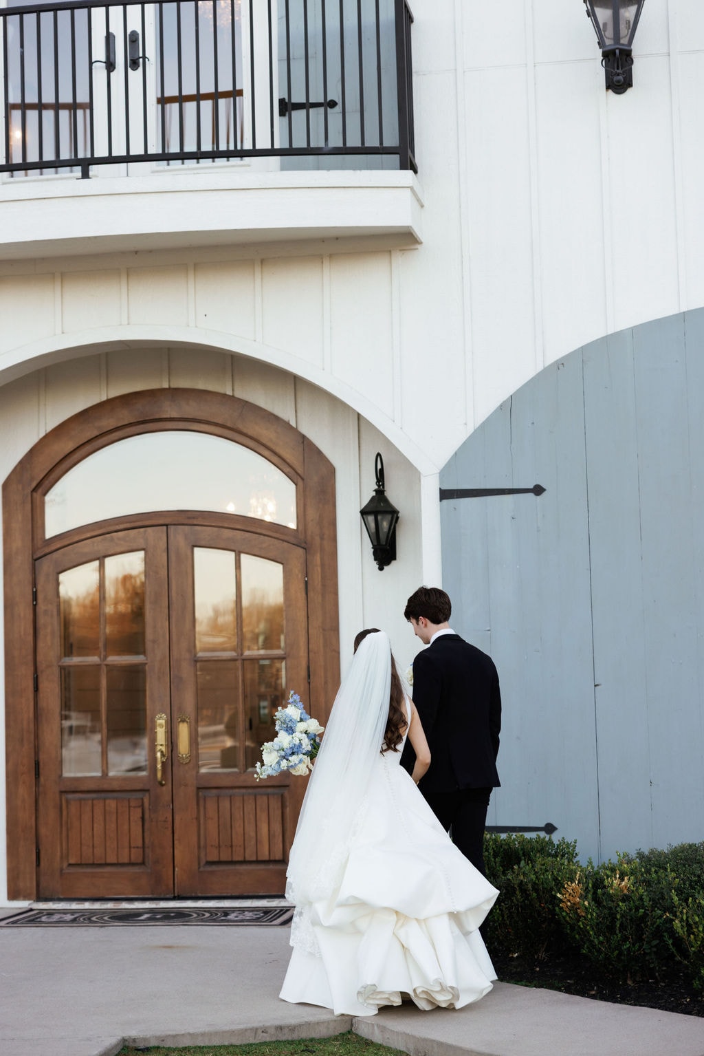 Brooke and Shepard walking hand in hand toward the arched wood double doors of the French Farmhouse venue, Brooke's cathedral veil and layered ballgown trailing behind her, the warm glow of the chandelier visible through the glass