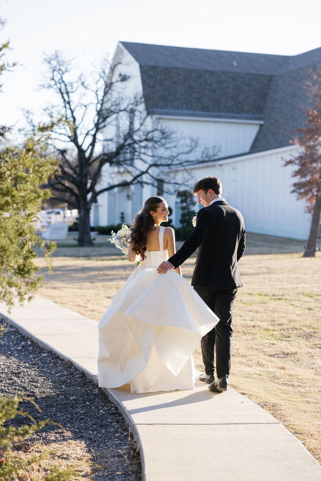 Shepard carrying Brooke's gown train as they walk the sunlit pathway on the French Farmhouse grounds, both laughing and looking at each other, the white barn and bare winter oak trees behind them