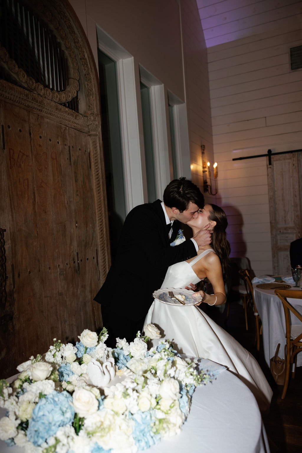 Brooke and Shepard sharing a kiss after cutting their cake in front of the antique carved wood barn doors inside The French Farmhouse reception hall, Brooke holding a plate of cake, blue and white floral arrangements on the cake table in the foreground, cross-back chairs and white shiplap walls glowing behind them