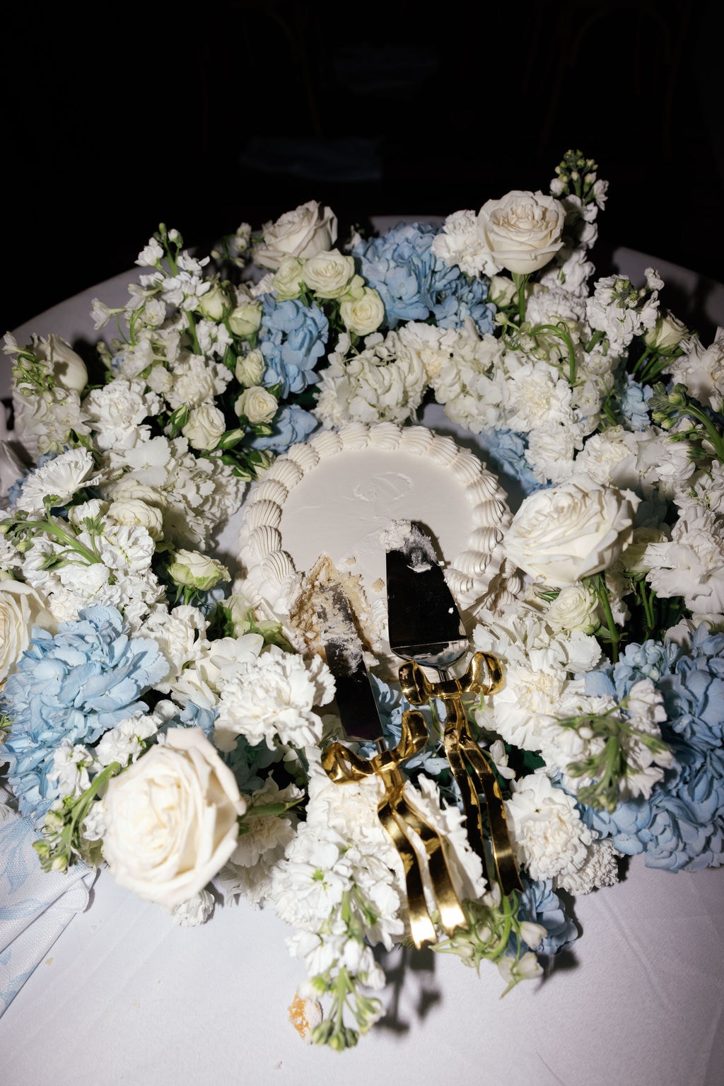 Close-up overhead shot of Brooke and Shepard's single-tier white wedding cake completely surrounded by a lush wreath of blue hydrangeas, white roses, white stock flowers, and spray roses on a white linen table, gold cake knife and server resting beside the first cut slice