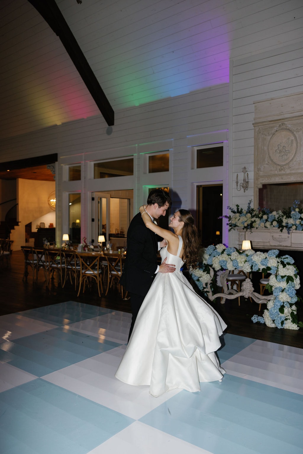 Brooke and Shepard swaying together during their first dance on the blue and white checkerboard dance floor inside The French Farmhouse reception hall, rainbow uplighting washing the white shiplap walls in color, blue hydrangea arrangements overflowing around the ornate French fireplace behind them, long farm tables with cross-back chairs glowing in the background