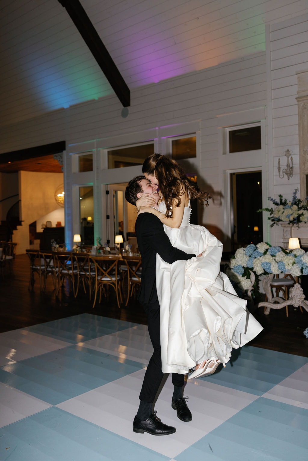 Shepard lifting Brooke off the ground during their first dance, both laughing and beaming, her white ballgown billowing around them on the blue and white checkerboard dance floor, rainbow uplighting and blue hydrangea fireplace arrangements glowing behind them