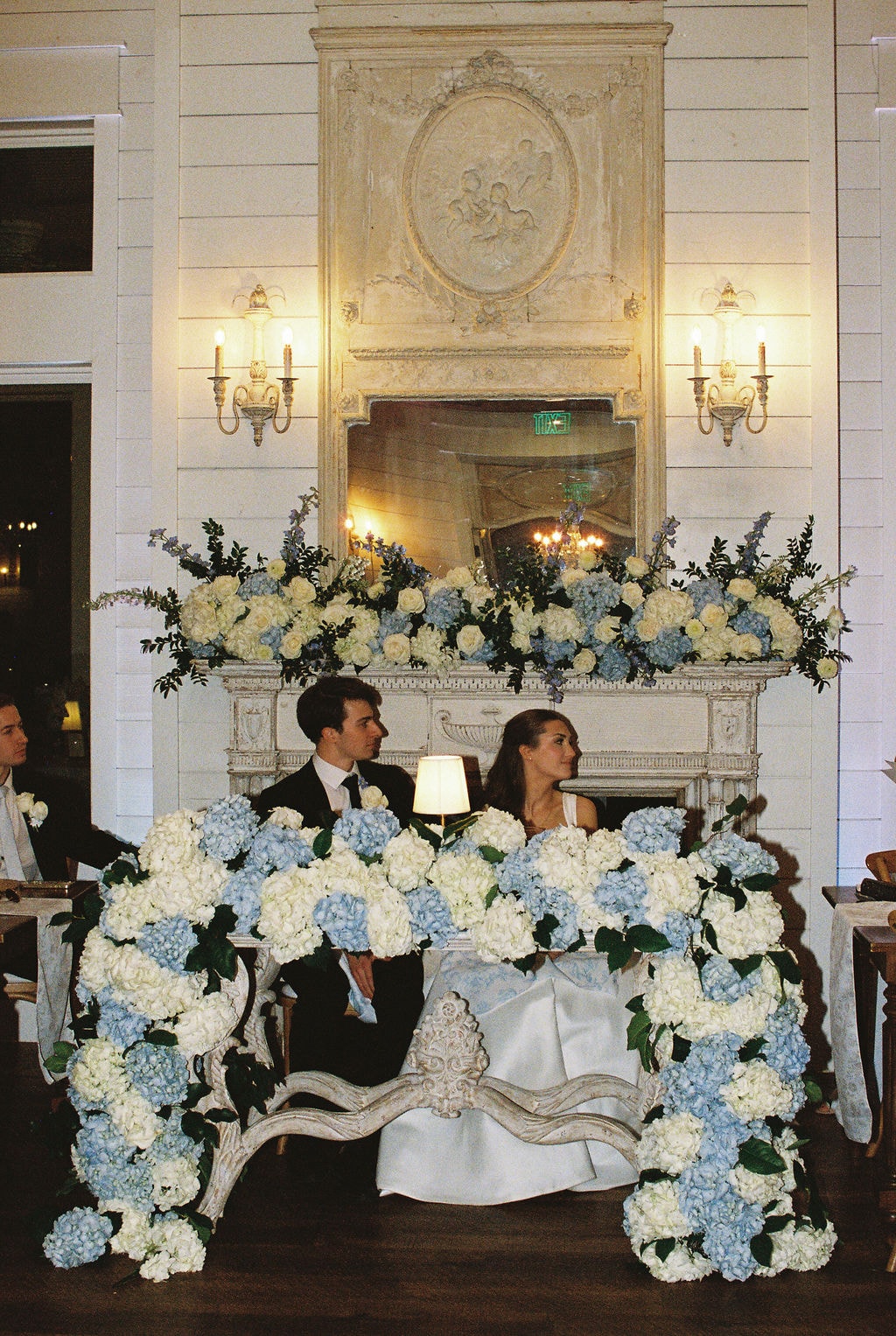 Brooke and Shepard seated at their sweetheart table in front of the ornate antique French fireplace inside The French Farmhouse reception hall, the table and loveseat completely covered in overflowing blue hydrangeas and white roses, matching floral garland cascading across the fireplace mantle above them, gold candelabra wall sconces glowing on either side