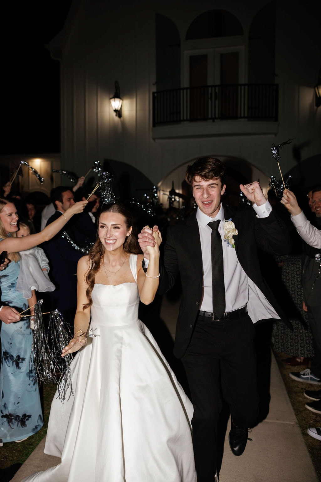 Brooke and Shepard running through a sparkler tunnel of cheering guests in front of the illuminated white French Farmhouse facade at night, Brooke's arms raised in pure joy, the arched entryway and wrought iron balcony glowing behind them