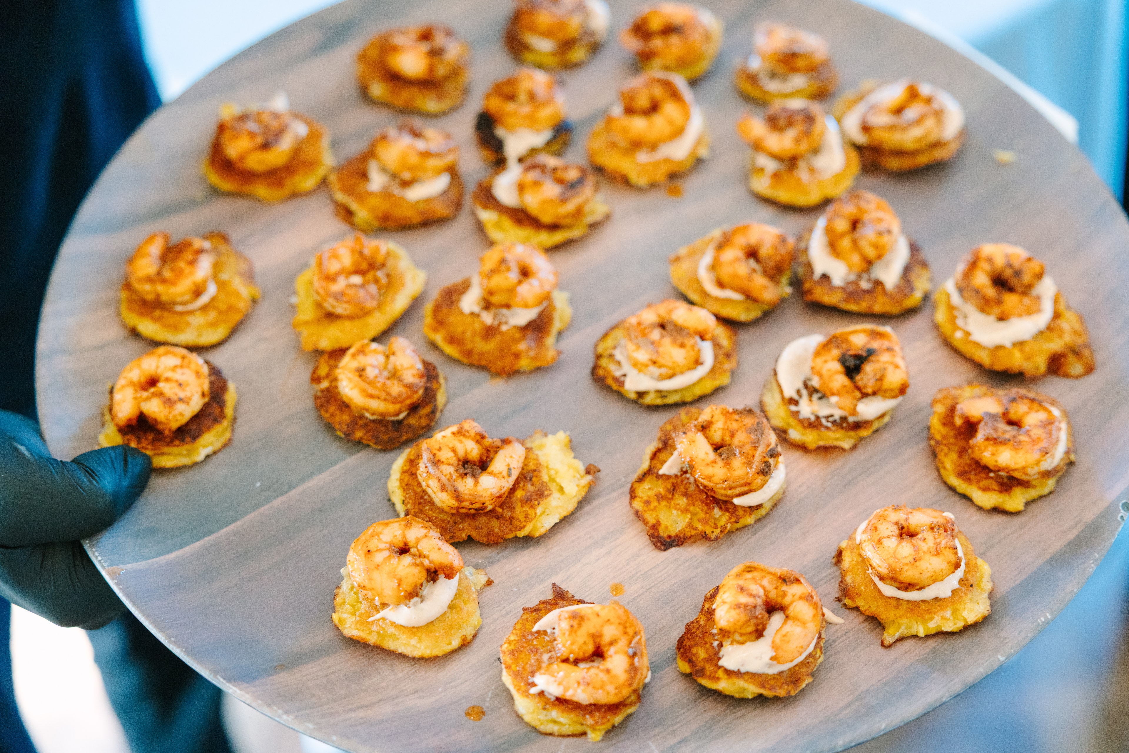 A round wooden board of passed appetizers — seasoned shrimp on golden polenta cakes with a dollop of cream sauce, held by a gloved server at a French Farmhouse Venue wedding