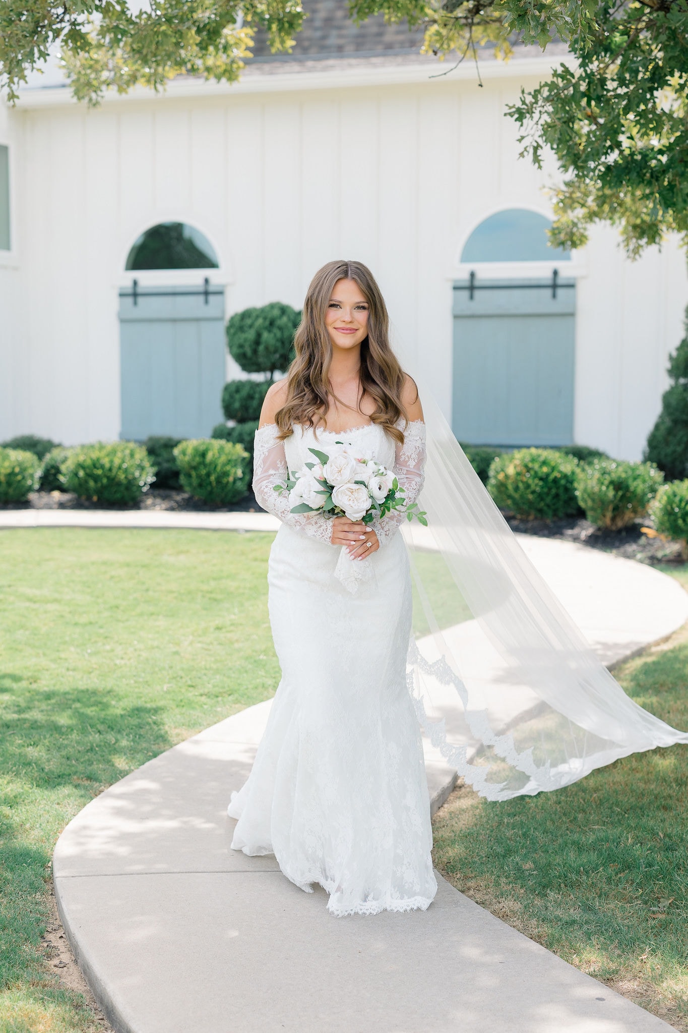 Catherine in her off-the-shoulder lace gown with veil flowing and white bouquet on the grounds of The French Farmhouse Venue