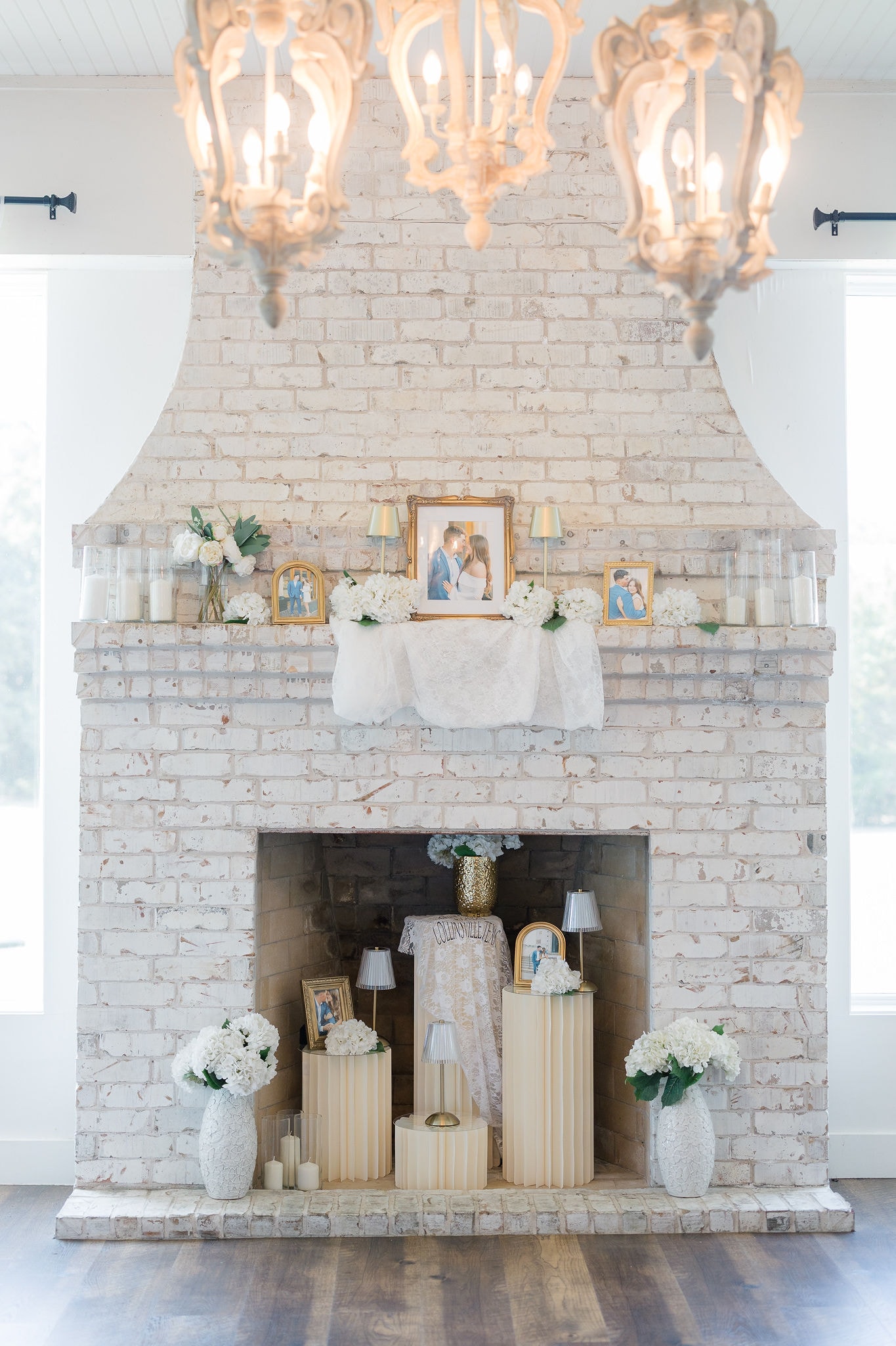 The French Farmhouse Venue's whitewashed brick fireplace styled with Catherine and Kade's engagement photos in gold frames, white hydrangeas, pillar candles, and a lace runner — crystal chandeliers overhead