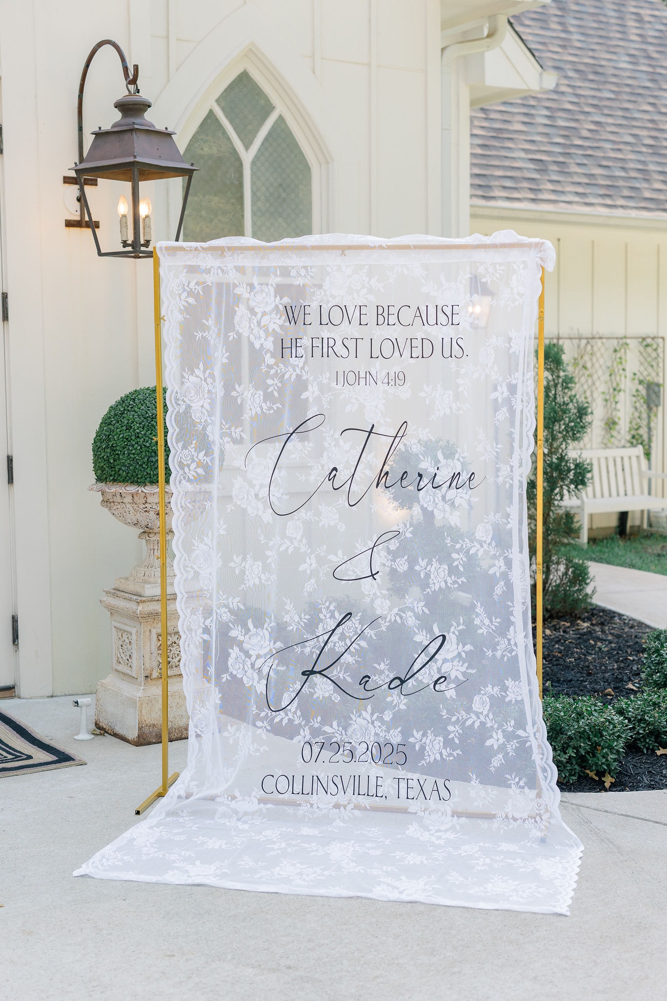 Catherine and Kade's lace wedding welcome sign reading 'We love because He first loved us — 1 John 4:19' with their names and date 07.25.2025, Collinsville Texas, displayed on a gold stand outside the chapel entrance