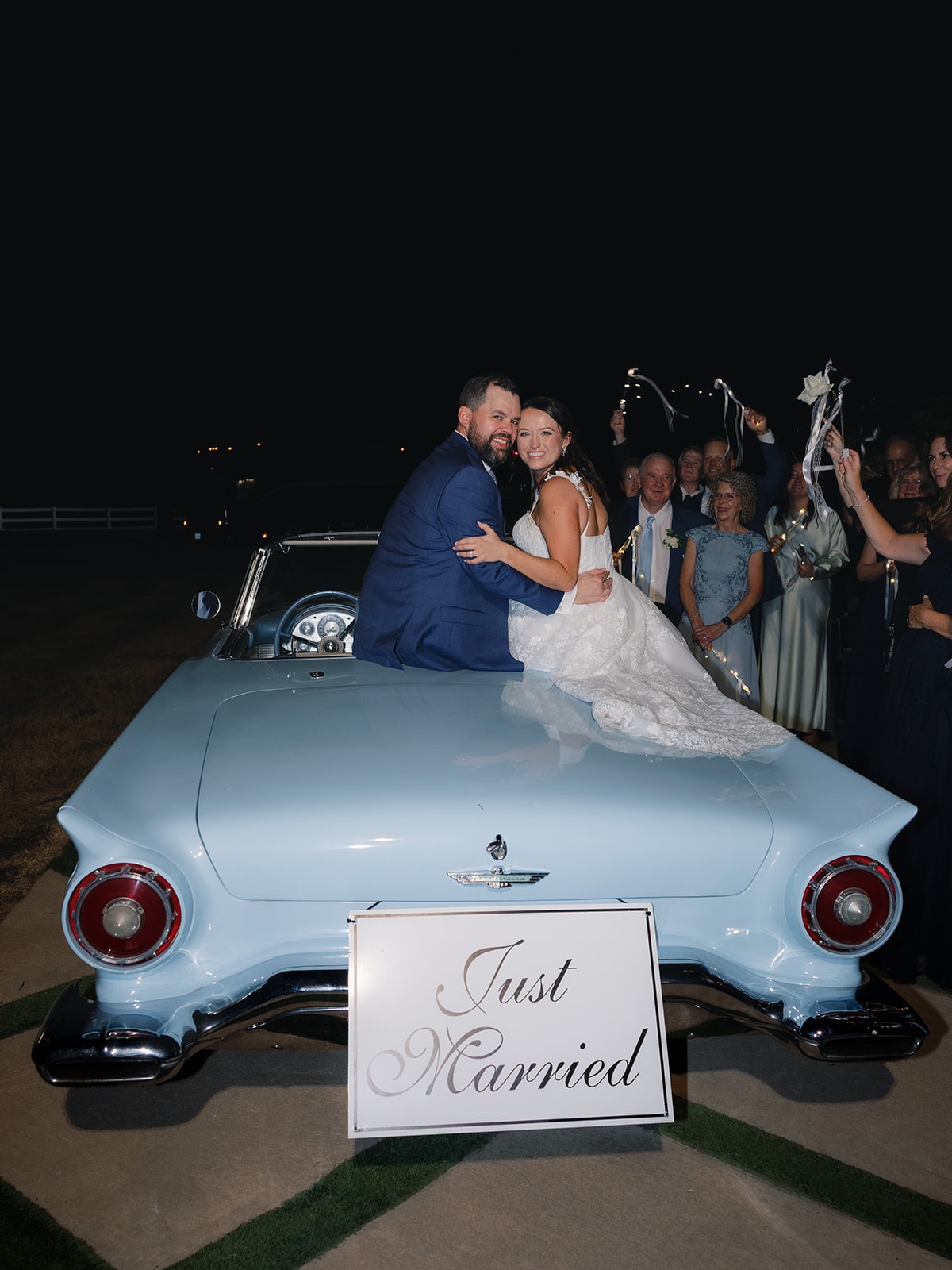 Corynn and CJ sitting on the back of a baby blue vintage Ford Thunderbird with a Just Married sign as guests wave ribbon wands