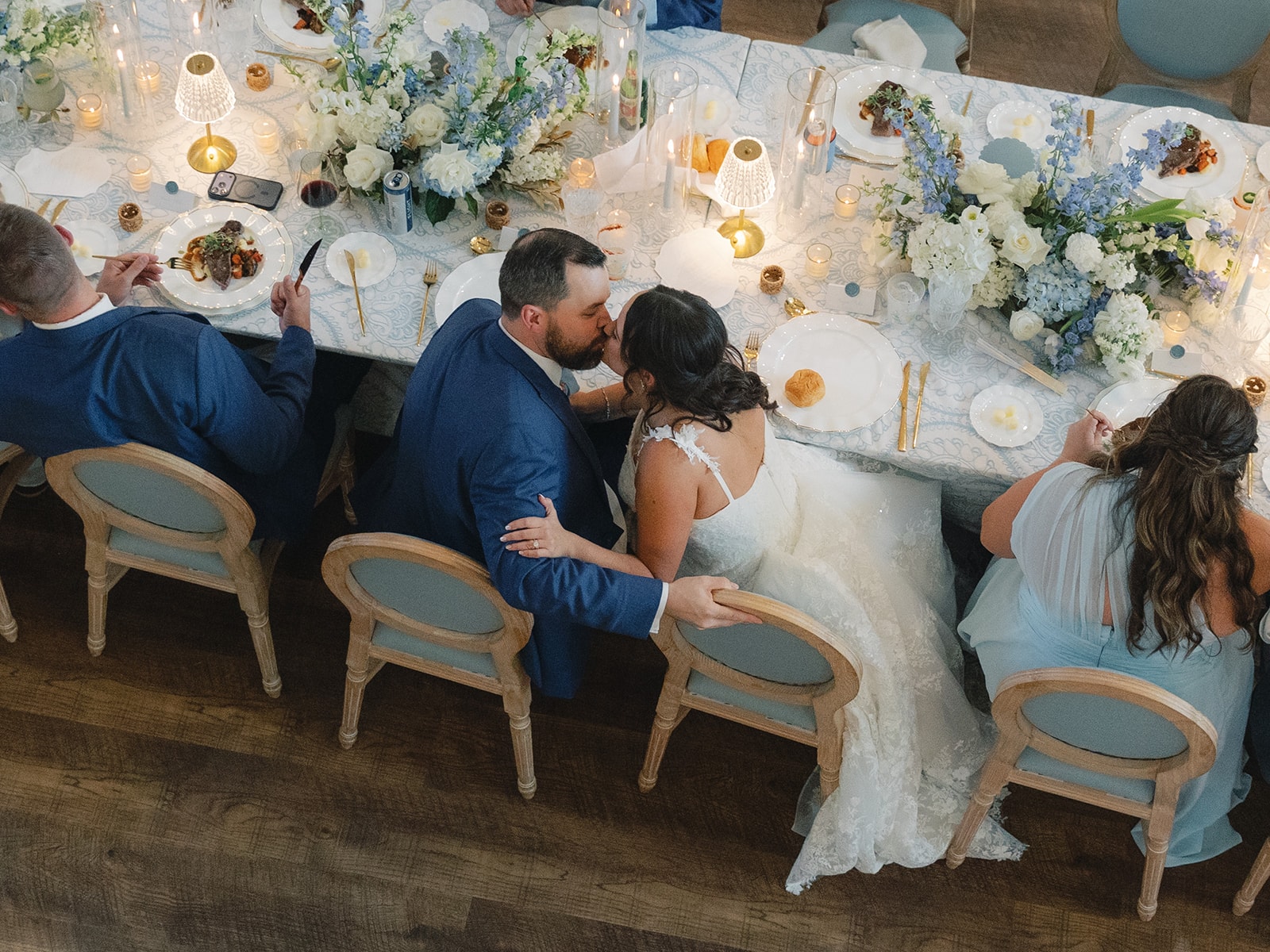 Aerial view of Corynn and CJ kissing at the sweetheart table surrounded by blue and white florals and candlelight