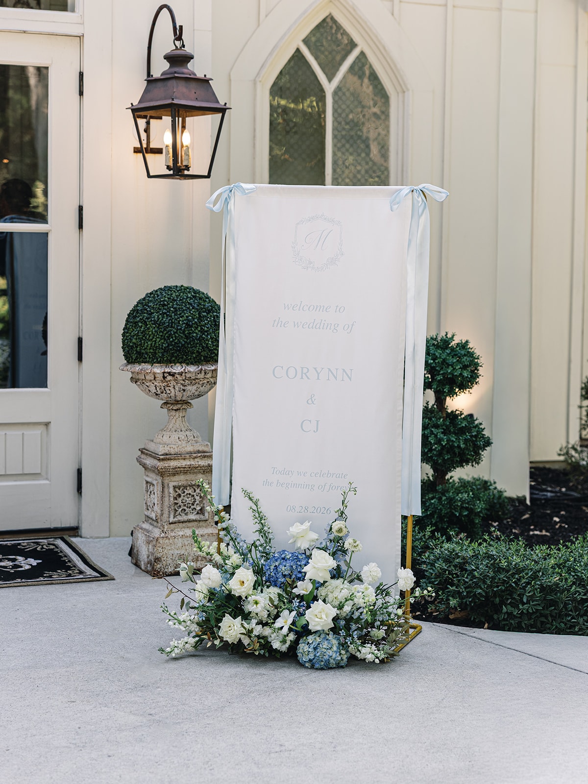 Welcome sign for Corynn and CJ's August 28, 2026 wedding with blue ribbon ties and white and blue florals at the base