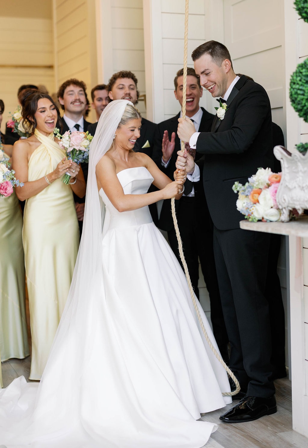Janie and Jake laughing together as they ring the chapel bell surrounded by their wedding party