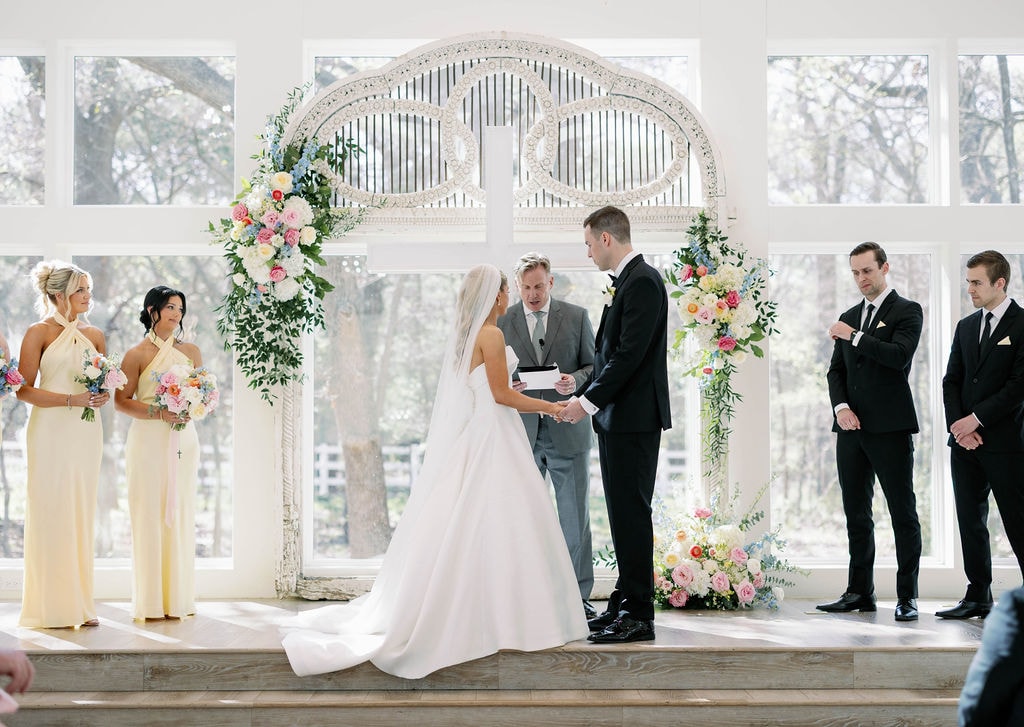 Wide shot of Janie and Jake at the altar with the full wedding party and colorful floral arch in the chapel