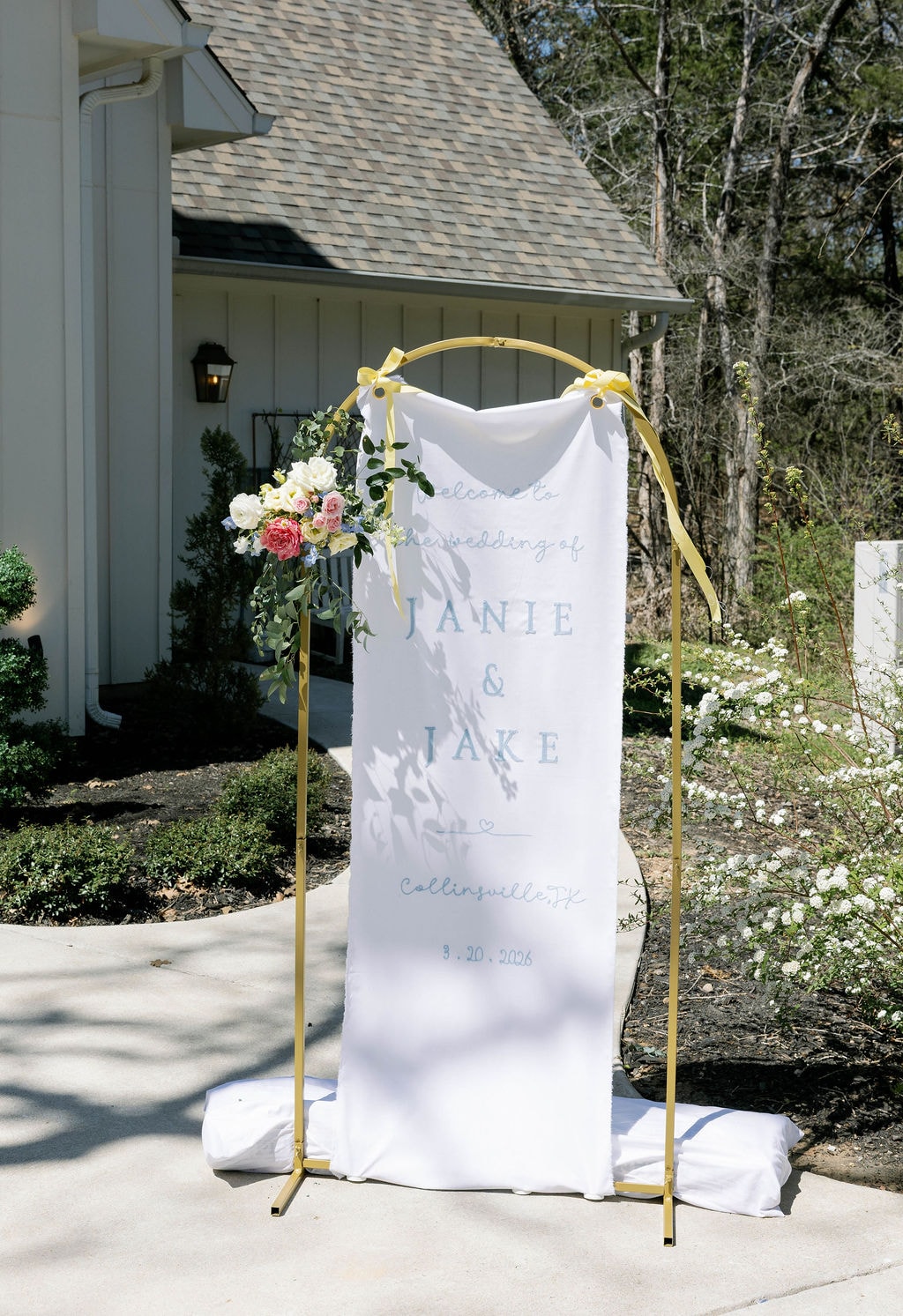 Janie and Jake welcome sign on gold arch with spring florals outside the chapel