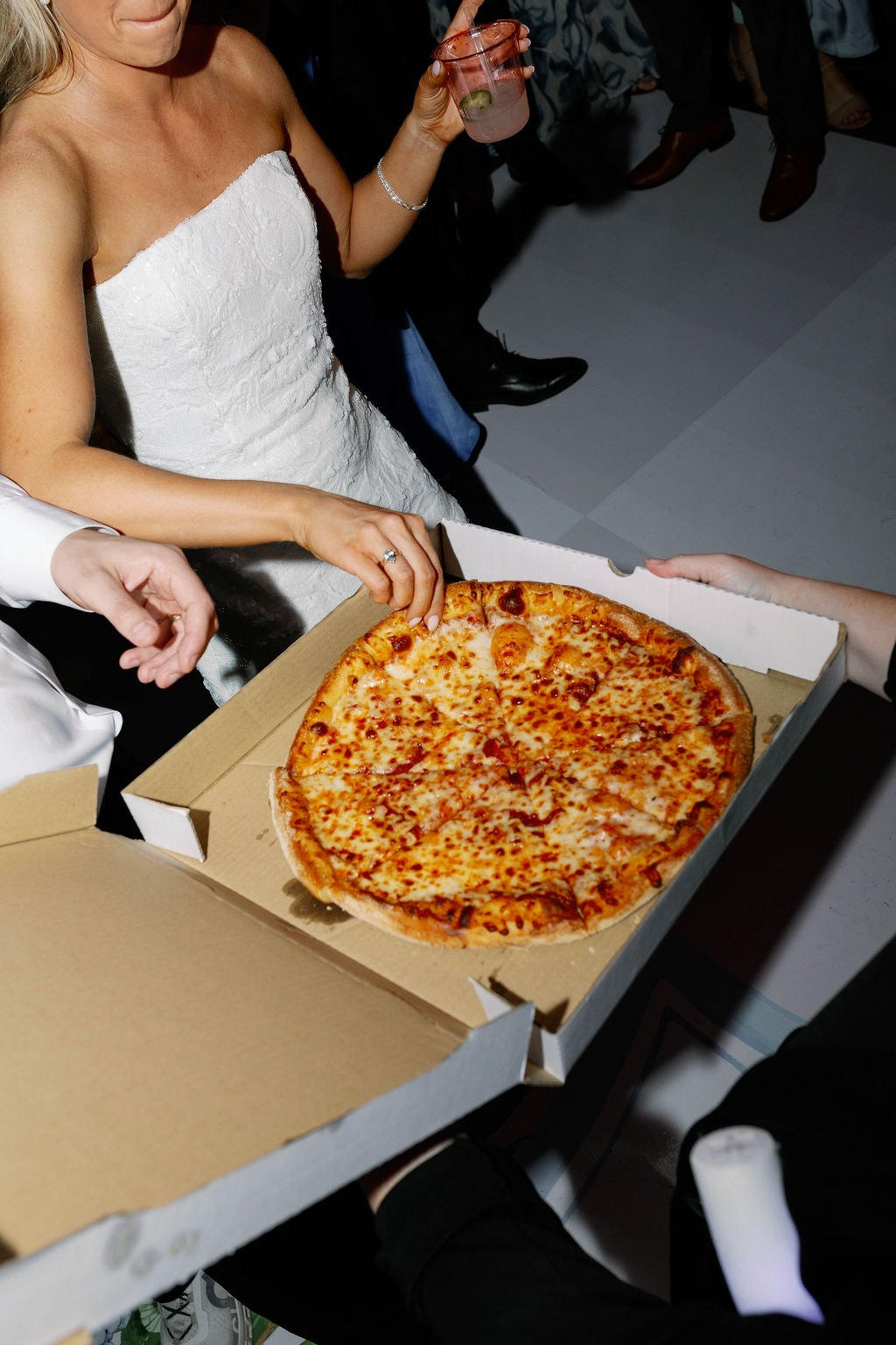 Bride in sequin mini dress grabbing a slice of pizza from a box on the dance floor late at night