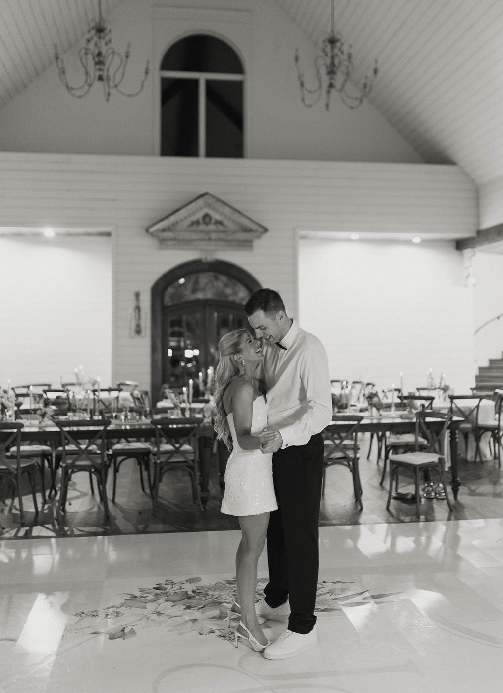Janie and Jake sharing a private last dance alone in the empty reception hall, chandeliers glowing above them, in black and white
