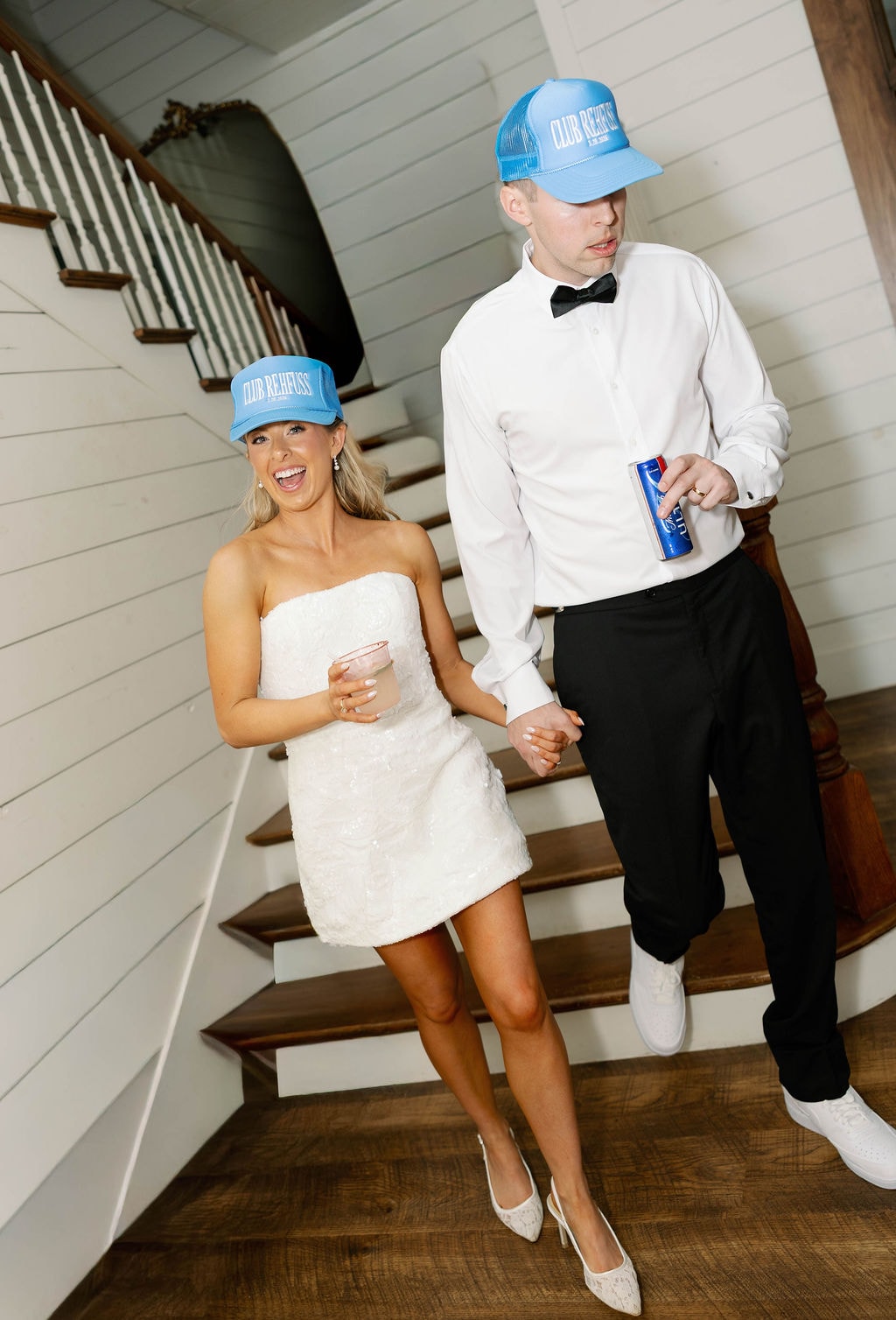 Janie in her reception mini dress and Jake in his untucked shirt, both wearing Club Rehfuss trucker hats on the farmhouse staircase
