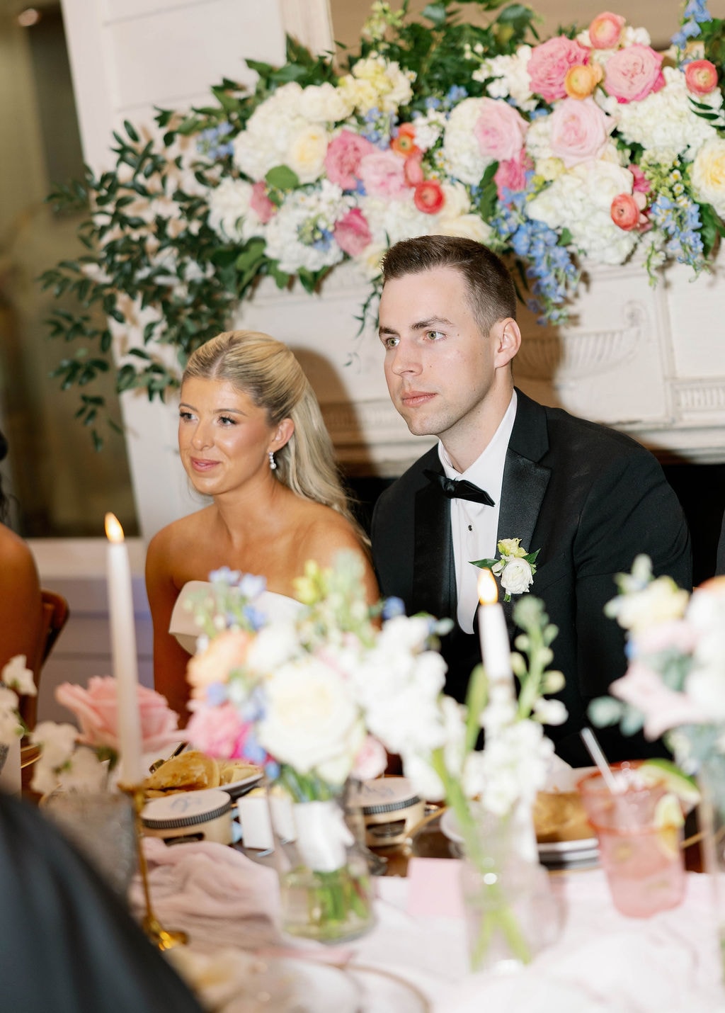 Janie and Jake at the sweetheart table listening to toasts, surrounded by lush pink and blue florals at the fireplace