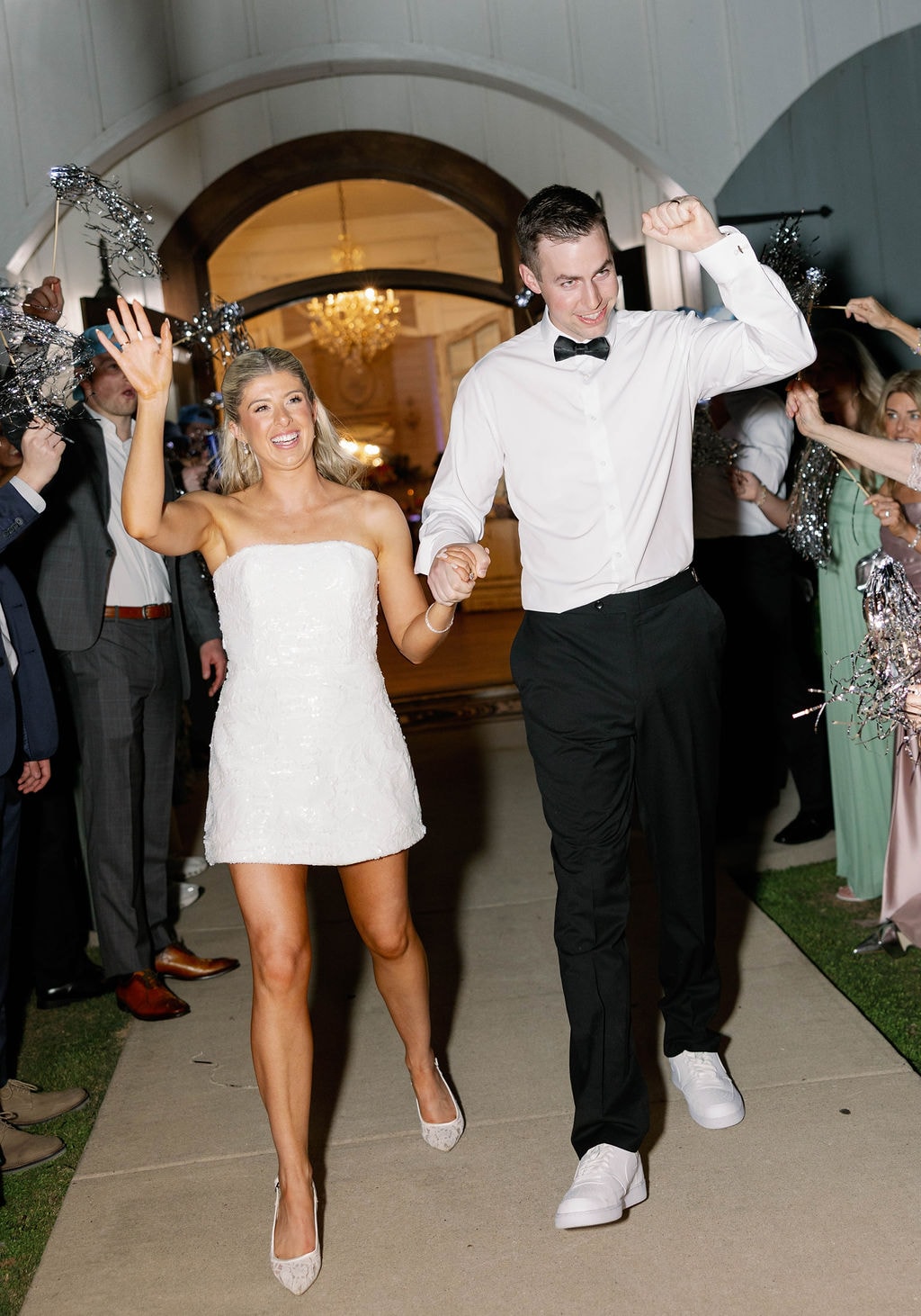 Janie and Jake bursting through the farmhouse doors during their send-off, guests lining the path with silver confetti wands, both laughing and hands raised