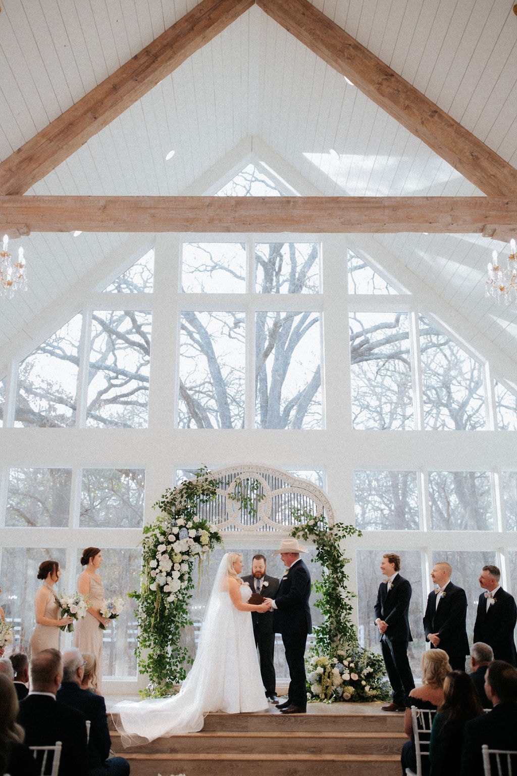 Wide shot of Kara and Anthony exchanging vows at the antique arch with lush white and green florals in the chapel