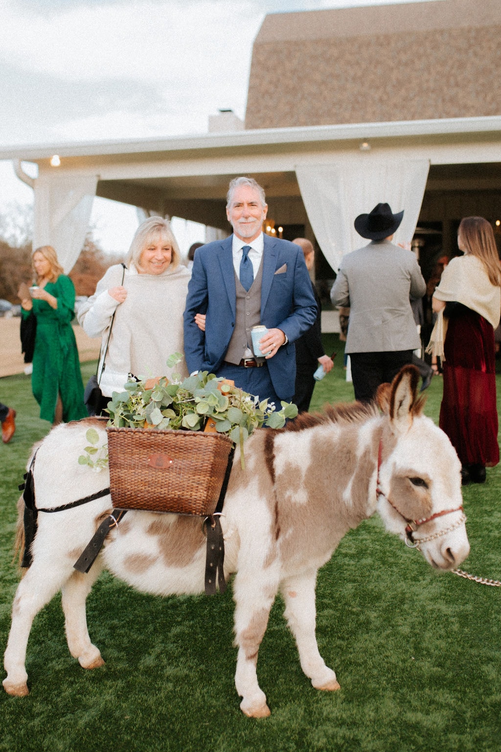 Wedding guests posing with a spotted mini donkey carrying a wicker basket of eucalyptus during cocktail hour on the lawn at The French Farmhouse Venue