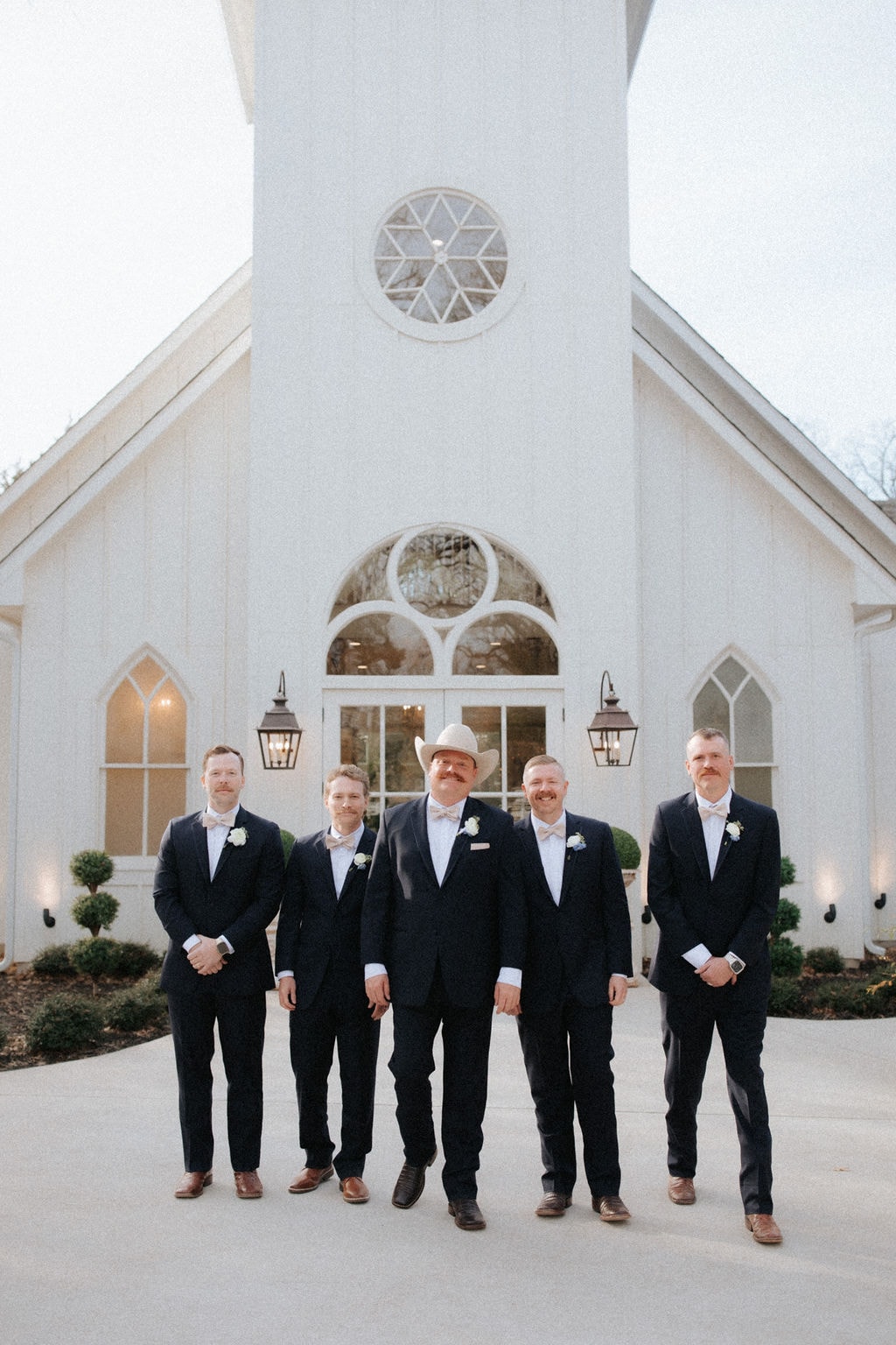 Anthony Marquardt and his four groomsmen in navy suits with blush bow ties standing in front of The French Farmhouse chapel at dusk — Anthony in the center wearing his cream cowboy hat