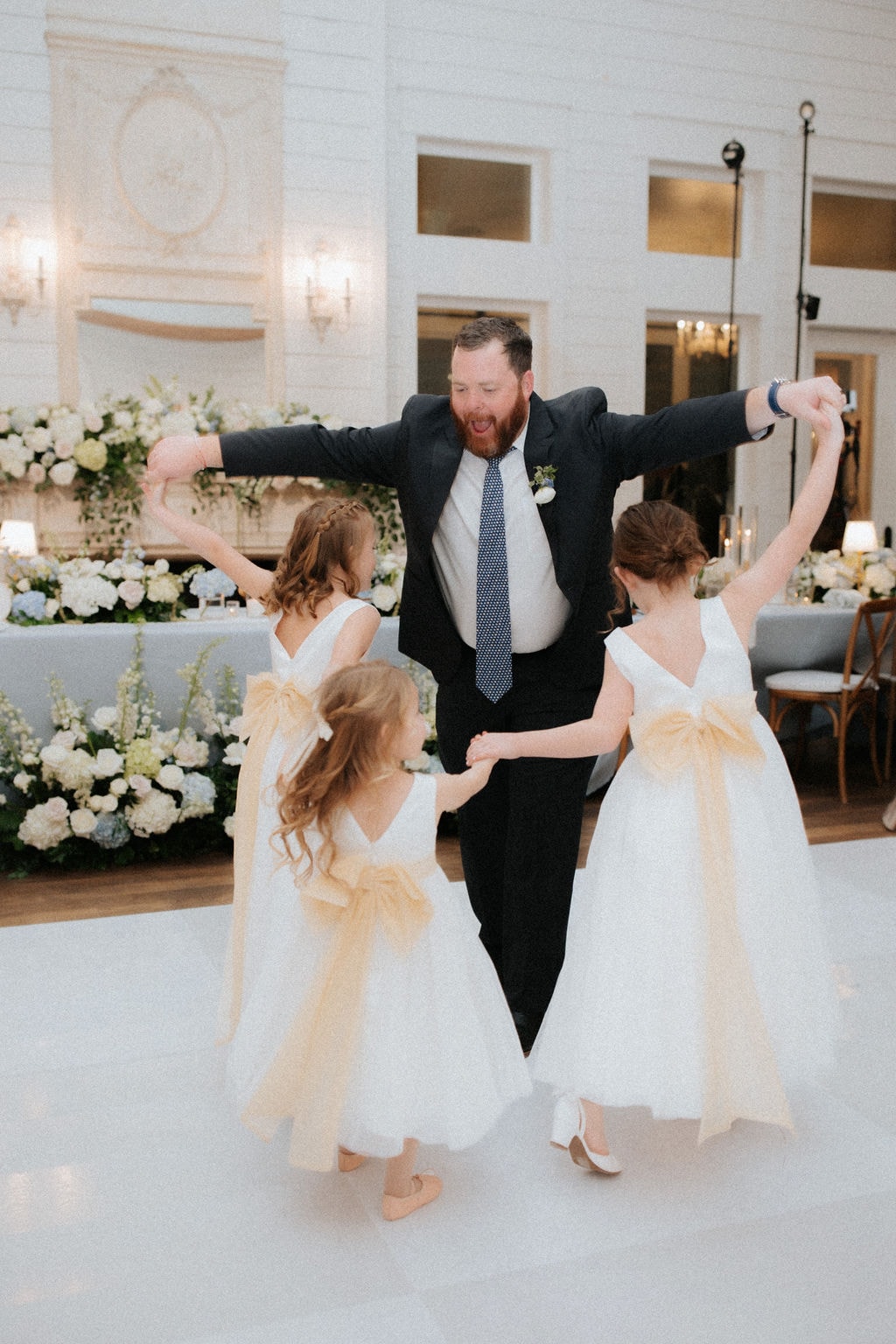 Anthony Marquardt dancing with three flower girls in white dresses with gold sashes on the dance floor at The French Farmhouse Venue reception, arms spread wide and laughing