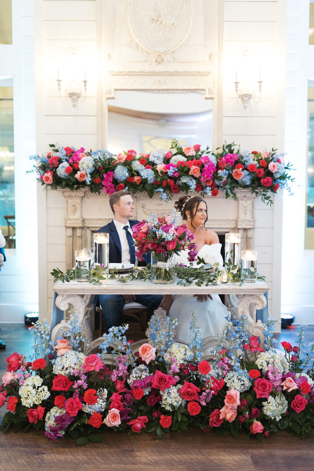 Katherine and Hayden at their sweetheart table surrounded by lush pink and blue florals at the fireplace